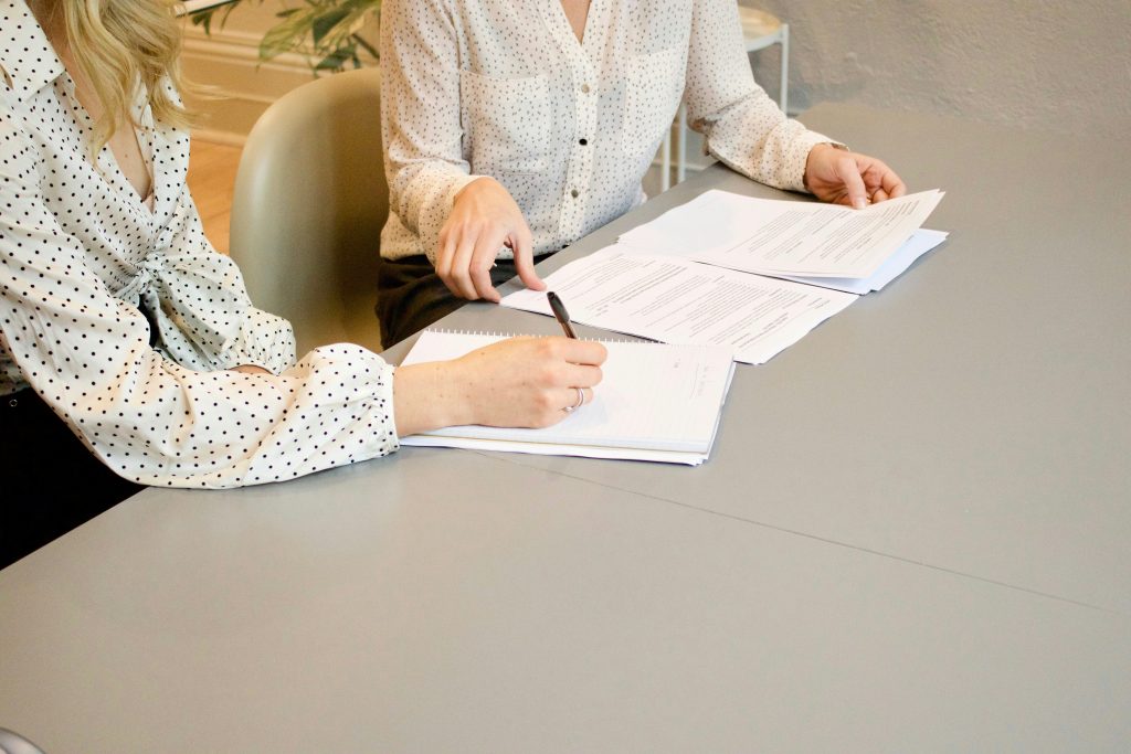 woman signing on white printer paper beside woman about to touch the documents woman signing on white printer paper beside woman about to touch the documents
